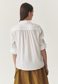 Witte blouse met een klassieke kraag, opgerolde mouwen en een geplooid detail aan de achterkant, gecombineerd met een beige rok. Soepele, lichte stof.