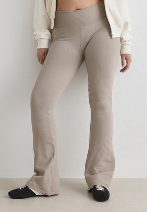 Kvinde iført beige, ribstrikkede bukser med høj talje og vide ben samt sorte og hvide sneakers, stående mod en ensfarvet væg.