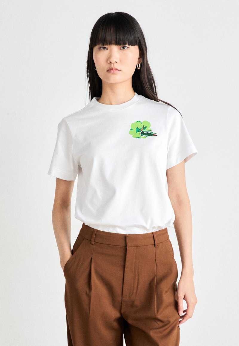 T-shirt bianco in cotone con un design grafico verde e nero sul lato sinistro del petto. La t-shirt ha maniche corte e colletto girocollo.