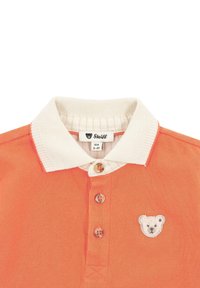 Oranžové polo tričko s bílým límcem, obsahující tři knoflíky a výšivku medvěda na levé hrudi. Texturovaná tkanina.