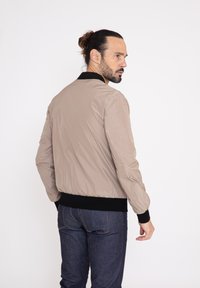Beige bomberjack met een zwarte geribbelde kraag en manchetten, met een gladde, glanzende textuur en elastische zoom, gedragen over donkere denim jeans.