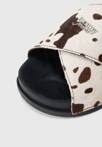 Σανδάλι slide με μοτίβο αγελαδινής γούνας, με ομαλή μαύρη λαστιχένια σόλα και μαλακό, επάνω μέρος από συνθ ετική γούνα με καφέ σημεία, διακοσμημένο με ασημένιο λογότυπο.