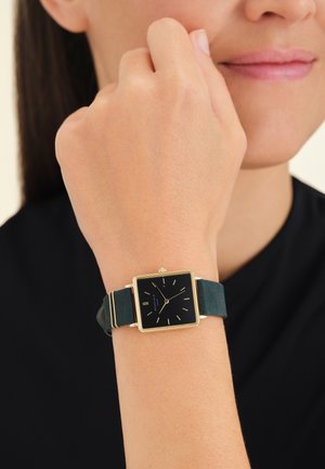 Montre carrée avec un cadran noir, des aiguilles dorées et un bracelet en suède vert. Boîtier carré avec des accents dorés. Portée au poignet.