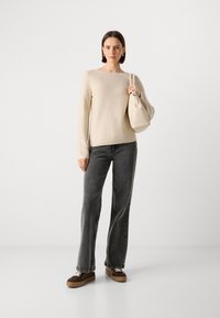 Suéter bege com um decote quadrado, combinado com jeans cinza escuro de cintura alta. Ela segura uma bolsa bege clara e usa ténis castanhos.