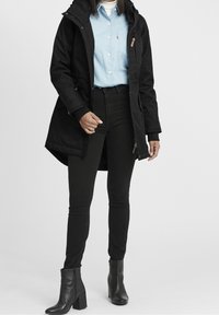 Svart lång kappa med huva, tillsammans med en ljusblå skjorta och högmidjade svarta jeans. Modellen bär svarta ankelstövlar.