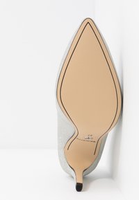 Chaussure à talon haut argentée à bout pointu positionnée semelle vers le haut, montrant une semelle beige avec contour noir et indication de la taille 37.