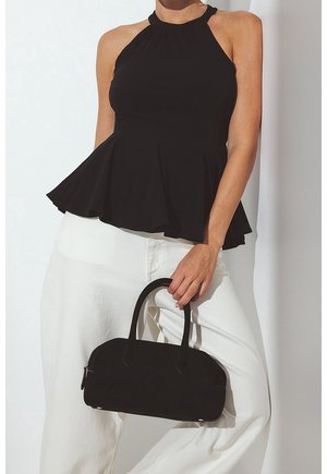 Kvinde iført sort ærmeløs peplumtop og hvide bukser, der holder en lille sort håndtaske i håndtagene i taljehøjde.