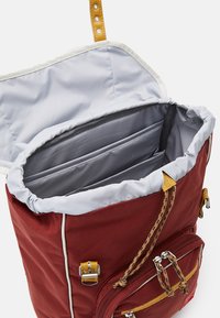 Sac à dos en toile rouge avec une doublure grise, des bretelles en cuir réglables et plusieurs poches intérieures. Dispose d'une poche zippée à l'avant et de quincaillerie en métal.