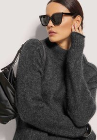 Donkergrijze, pluizige gebreide trui met een hoge halslijn en lange mouwen. Gecombineerd met zwarte cat-eye zonnebril en een zwarte tas met een kettingriem.