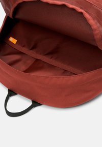 Röd tygryggsäck med ett svart handtag, öppen insida och en enda orange varumärkesetikett. Har en slät textur och rund form.