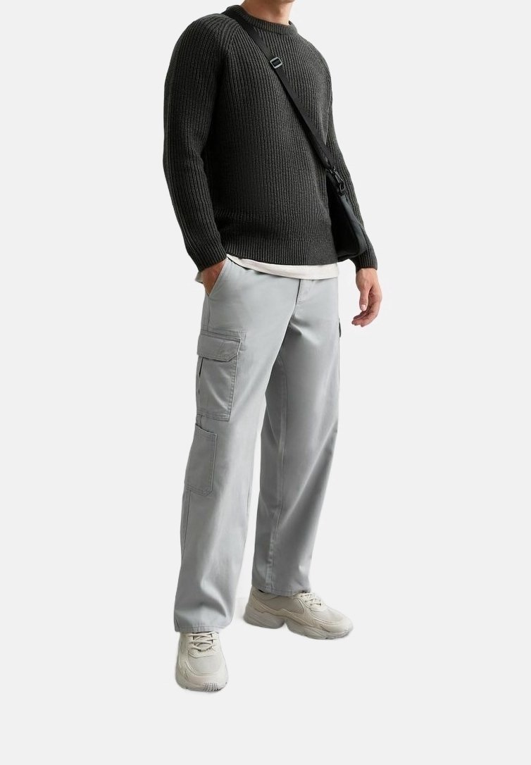 Pantalon cargo gris avec poches latérales, associé à un pull côtelé foncé et un t-shirt clair. La tenue est complétée par des baskets beige et un sac noir.