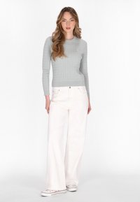 Maglione a costine grigio chiaro con maniche lunghe, abbinato a pantaloni bianchi a gamba larga e sneakers bianche, in piedi davanti a uno sfondo neutro.