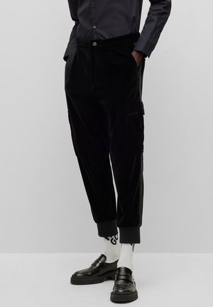 Mies, jolla on mustat cargo-housut, valkoiset sukat mustalla tekstillä, mustat loafers-kengät ja tumma napillinen paita, kädet taskuissa.