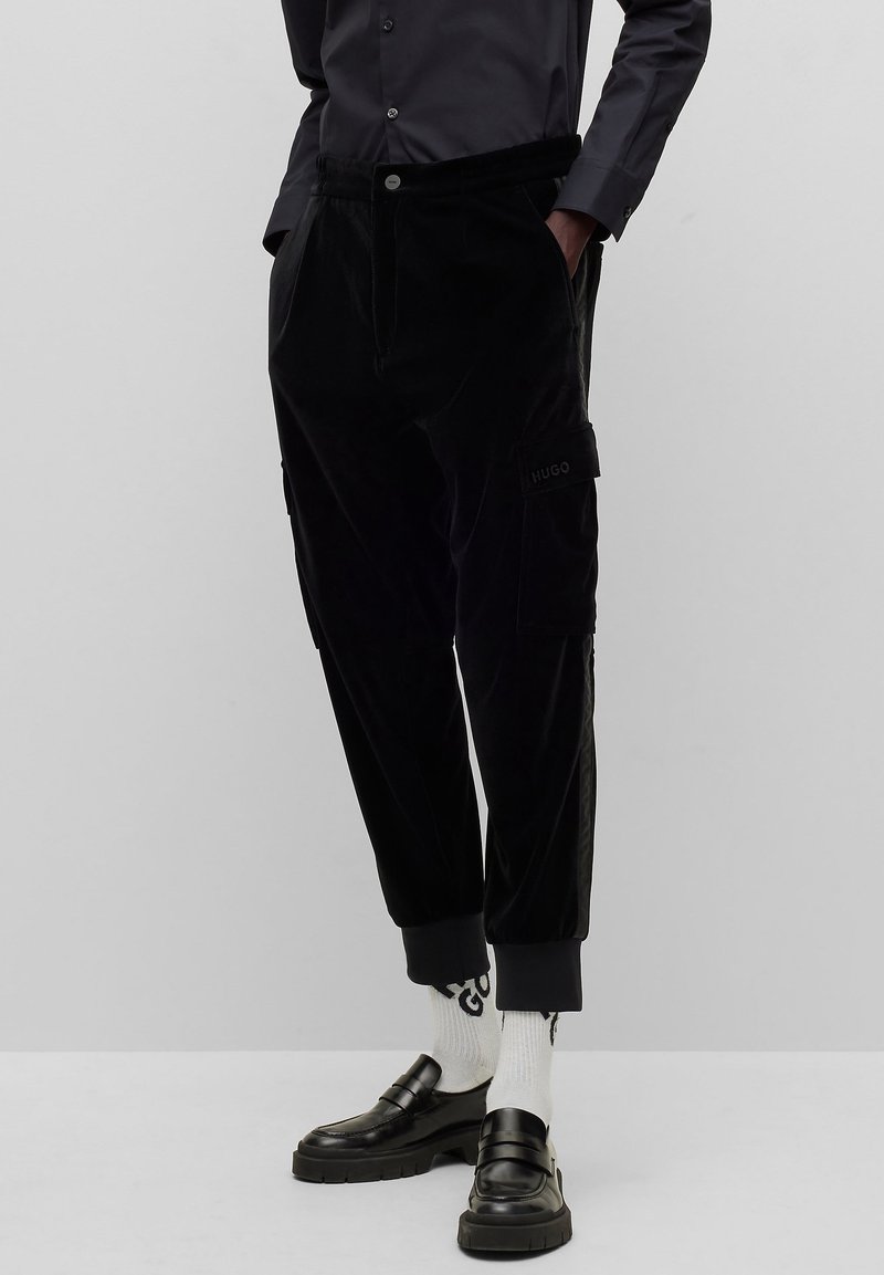 Homme portant un pantalon cargo noir, des chaussettes blanches avec des lettres noires, des mocassins noirs et une chemise sombre boutonnée, les mains dans les poches.