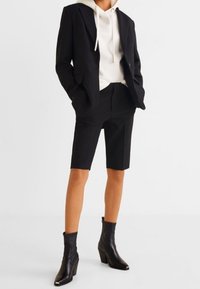 Blazer negro entallado sobre una sudadera crema, combinado con pantalones cortos negros y botas al tobillo. El conjunto presenta líneas limpias y formas estructuradas.
