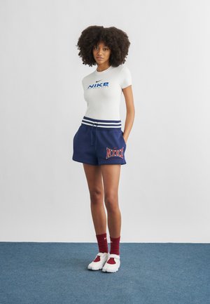 Kobieta o kręconych włosach w białej koszulce z logo Nike, granatowych szortach z nadrukiem Nike, czerwonych skarpetkach i białych butach stojąca na niebieskim dywanie.