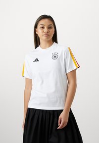 Λευκό κοντομάνικο μπλουζάκι με μαύρο λογότυπο Adidas και έμβλημα ποδοσφαίρου Γερμανίας. Διαθέτει κόκκινες, κίτρινες και μαύρες ρίγες στα μανίκια. Κατασκευασμένο από βαμβακερό υλικό.