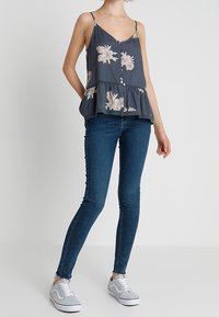 Dark gray floral tank top with adjustable straps, gathered waist, and button front. Paired with slim blue jeans and gray sneakers.