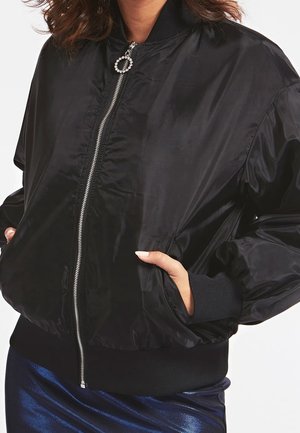 Kobieta nosi czarną satynową kurtkę bomber z srebrnym zamkiem błyskawicznym i okrągłym suwakiem z cyrkoniami, ręce w bocznych kieszeniach, w połączeniu z ciemnoniebieską spódnicą.