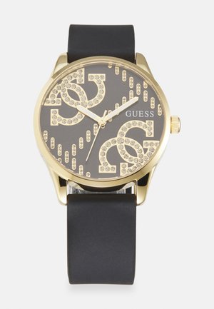 Montre dorée avec un bracelet en caoutchouc noir. Le cadran analogique présente des lettres métalliques et des cristaux sur un fond sombre.