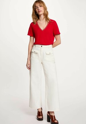 Mujer con camiseta roja de manga corta y escote en V, pantalones blancos de pierna ancha, de talle alto y con bolsillos delanteros, y sandalias marrones con tacón ancho.