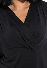 Mustassa kietomopaidassa on V-pääntie, pehmeä kangas ja hienovaraiset poimutustiedot, sekä sileä pinta ja vartalonmyötäinen siluetti.