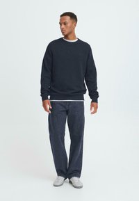 Marineblauer Rundhalsausschnitt-Sweatshirt aus Strickstoff, über einem weißen T-Shirt getragen. Kombiniert mit dunklen, schmal zulaufenden Jeans und hellgrauen Sneakers.
