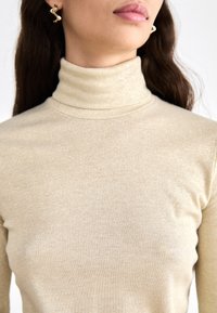 Beige ribattu neulepaita, jossa on hienovarainen kiilto. Mallissa on säädettävä siluetti ja korkea, jämäkkä kaulus. Kultaiset kiehkura-korvakorut.