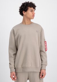 Felpa beige realizzata in tessuto morbido, presenta polsini e orlo a coste, tasca con zip sul braccio sinistro e un dettaglio di etichetta rossa appesa.