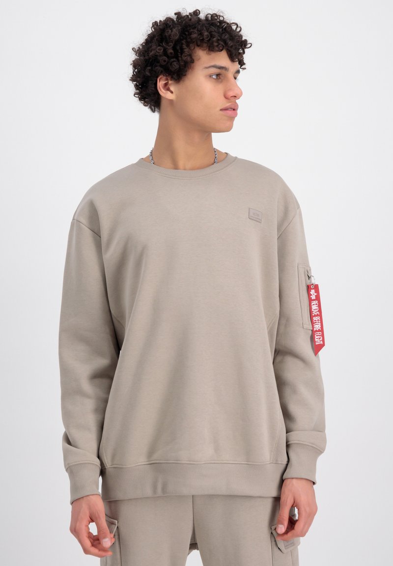 Felpa beige realizzata in tessuto morbido, presenta polsini e orlo a coste, tasca con zip sul braccio sinistro e un dettaglio di etichetta rossa appesa.