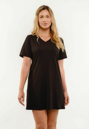 Lyshåret kvinde iført en enkel sort kortærmet V-hals kjole, stående mod hvid baggrund, vendt fremad med afslappet kropssprog.