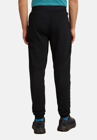 Ein Mann trägt schwarze tapered Hose, blaue Socken und schwarze Wanderschuhe, von hinten vor einem schlichten weißen Hintergrund gezeigt.