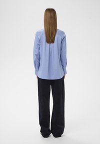 Chemise bleu clair à boutons avec manches longues, détail de pli dans le dos, associée à un pantalon ample bleu marine foncé en tissu lisse.