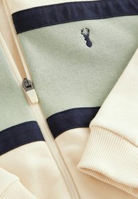 Svetlo béžová bundu na zips so zelenými a námorníckymi horizontálnymi pruhmi, ribbed manžetami a malým vyšívaným logom na hrudi. Mäkká textúra látky.