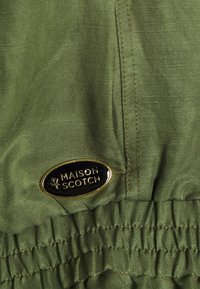 Tissu vert olive avec une texture lisse, présentant une taille élastique froncée. Un patch rond noir et or affiche "MAISON SCOTCH".