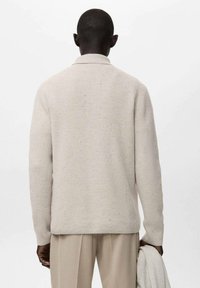 Jasnobeżowy, dziany sweter o luźnym kroju, z prążkowaną fakturą i kołnierzem. Modelka trzyma pasujący sweter.