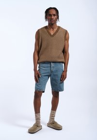 Maglione senza maniche marrone texturizzato, pantaloni shorts in denim blu, scarpe slip-on beige e calze lavorate marroni indossati da una persona che sta in piedi contro uno sfondo neutro.