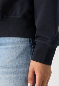 Main reposant sur le côté, portant un haut noir à manches longues avec poignets et un jean en denim bleu clair sur un fond uni.