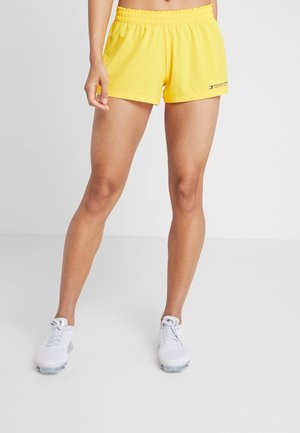 Person iført gule atletikshorts med elastisk talje og hvide sneakers, stående på en ensfarvet lys baggrund.