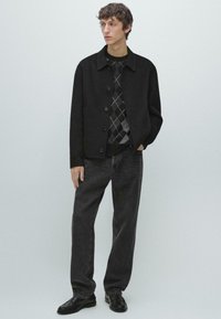 Sort jakke i uldblanding med knaplukning, båret over en mønstret sort og grå sweater. Kombineret med mørke jeans og sorte sko.