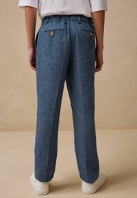 Blauwe textuurbroek met een relaxed fit, voorzien van een elastische tailleband, twee achterzakken met knopen en taps toelopende pijpen. Witte sneakers.
