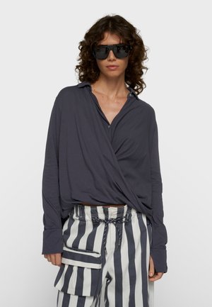 Mujer con cabello rizado que lleva gafas de sol negras de gran tamaño, blusa envolvente suelta de color gris oscuro y pantalones a rayas de talle alto con bolsillo lateral.