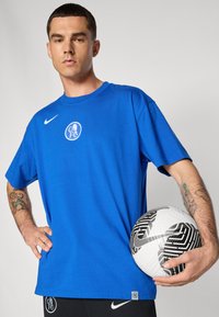 Blå bomulds t-shirt med rund halsudskæring, med et hvidt logo på brystet. Modellen holder en sort-hvid mønstret fodbold.