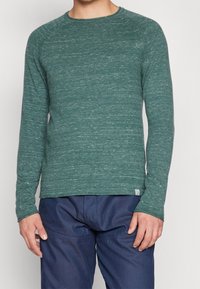 Långärmad grön skjorta i texturerad tyg, med rund halsringning och subtilt prickigt mönster. Matchas med mörkblå byxor.