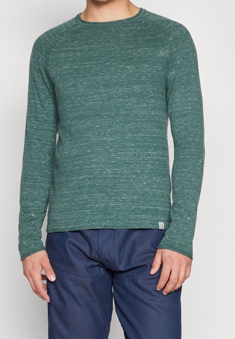 Långärmad grön skjorta i texturerad tyg, med rund halsringning och subtilt prickigt mönster. Matchas med mörkblå byxor.