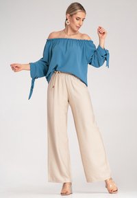 Jasnoniebieska bluzka typu off-the-shoulder z luźnymi rękawami i akcentami w postaci sznureczków, zestawiona z beżowymi spodniami o szerokich nogawkach, prezentującymi luźny krój.
