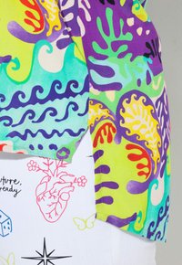 Multicolore korte mouwen shirt met abstracte vormen en golvende patronen in groen, paars en blauw; de stof lijkt glad en lichtgewicht.