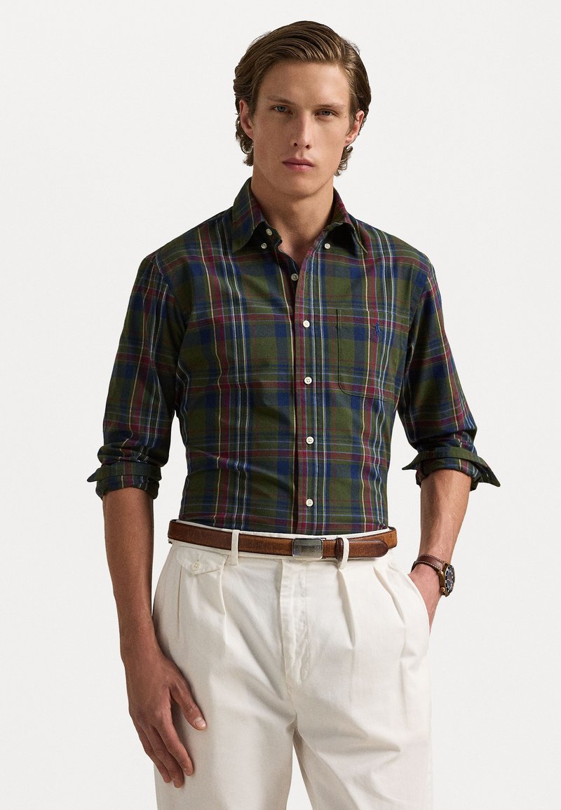 Groen geruite shirt met marineblauwe, rode en gele accenten, button-down kraag, opgestroopte mouwen, gecombineerd met crèmekleurige broek en een bruine riem.