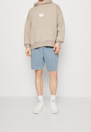 Person, die einen beigen oversized Hoodie, hellblaue Jeansshorts, weiße Socken und weiße Sneakers mit "Diesel"-Marke trägt.