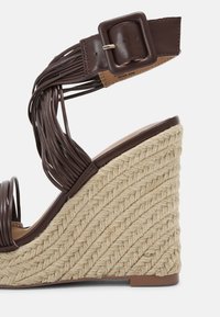 Sandale à talon compensé marron avec des sangles en synthétique tressées et une boucle ajustable. Dotée d'un talon enveloppé de jute texturé. Design à bout fermé.
